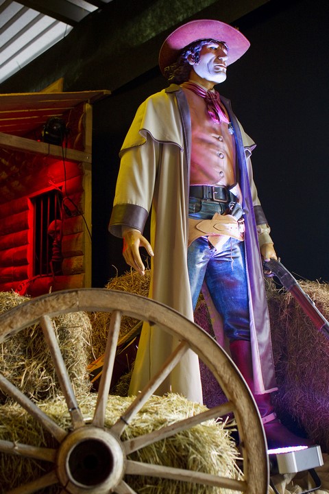 Statue grandeur nature d'un cow-boy en tenue western traditionnelle, posant à côté d'une roue de chariot et de bottes de paille, sous un éclairage scénique coloré, dans un décor de location pour événement à thème western
