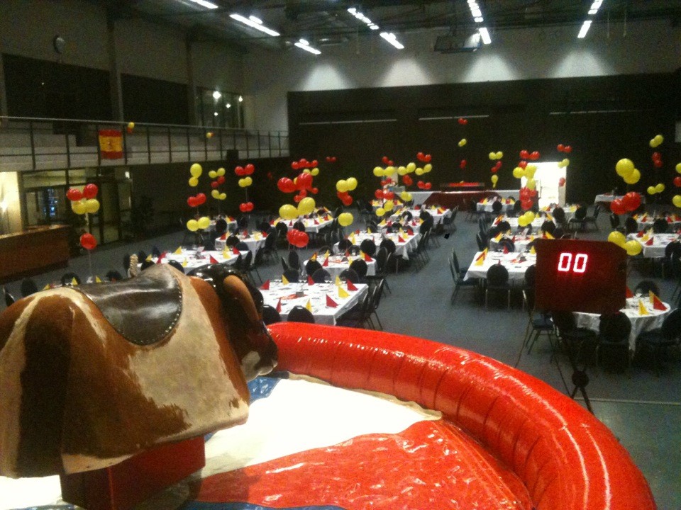 Vue sur un taureau mécanique au premier plan avec une salle de fête en arrière-plan, décorée de tables avec nappes, et ballons rouges et jaunes, prête pour une soirée à thème